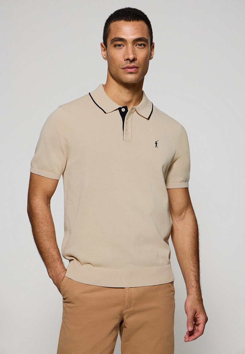 Polo shirt beige a maniche corte con colletto nero, dotato di chiusura frontale con bottoni e un piccolo logo ricamato sul lato sinistro del petto.