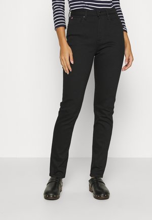 Persona que lleva unos jeans ajustados negros, zuecos negros con tachuelas y una camiseta de rayas azul marino y blanca de manga larga, de pie frente a un fondo blanco.