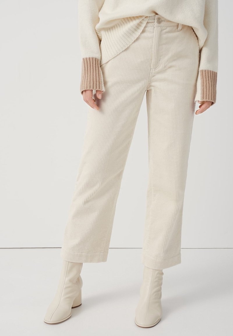 Femme portant un pantalon en velours côtelé crème, un pull crème avec des manches beiges et des bottines à talons crème sur un fond blanc.
