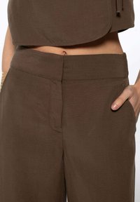 Pantalon marron taille haute avec une fermeture frontale dissimulée, porté avec un crop top assorti, main dans la poche droite, bracelets en or au poignet gauche.