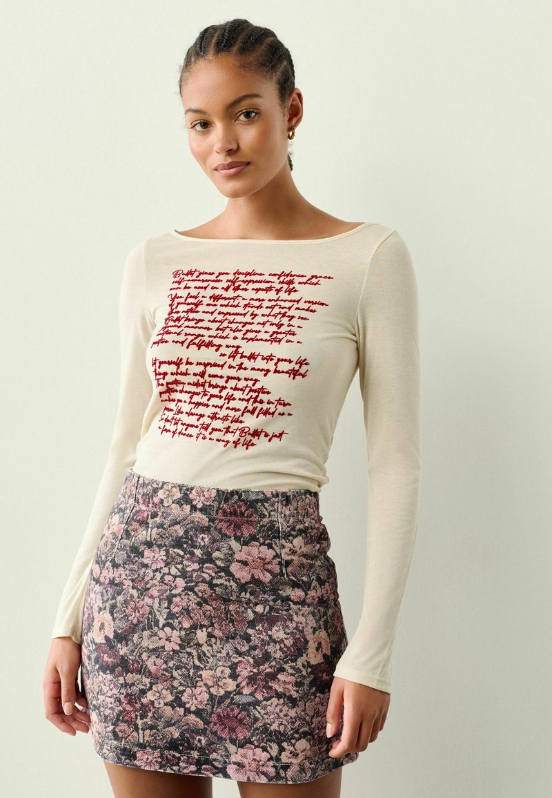 Langærmet creme top med rødt trykt tekst, parret med en blomster mini-nederdel i nuancer af pink, sort og grøn med en struktureret overflade.