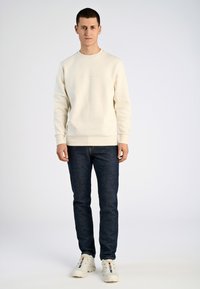 Krämfärgad sweatshirt med ribbstickade muddar och fåll, tillsammans med mörkblå jeans och vita sneakers. Modellen står mot en enfärgad bakgrund.