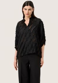 Blusa nera con tessuto trasparente e testurizzato, caratterizzata da una vestibilità comoda, scollatura a V e dettagli a righe sfrangiati. Abbinata a pantaloni neri.