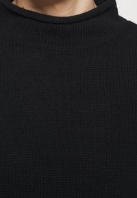 Gros plan d'une personne portant un pull en maille côtelée noire avec un col rond haut sur un fond uni.