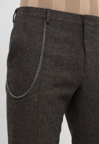 Donkergrijze, textuurgegeven wollen broek met een subtiel herringbonepatroon, voorzien van een zilveren ketting die ter decoratie is bevestigd.
