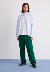 Lyseblå, stribet, skjorte med knapper over en hvid t-shirt, parret med grønne træningsbukser med hvide sidestriber og sorte sko.