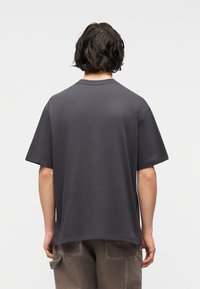 Άτομο με κοντά κυματιστά μαλλιά, γυρισμένο πλάτη, φοράει ένα φαρδύ σκούρο γκρι κοντομάνικο μπλουζάκι και καφέ παντελόνι με πίσω τσέπες.