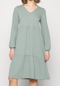 Robe verte claire avec décolleté en V, manches longues, motif zigzag texturé, et une jupe à volants avec des détails froncés aux poignets.