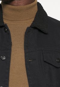 Musta farkkutakki, jossa on teksturoidusta mustasta sherpa-kankaasta valmistettu kaulus. Alla ruskea ribbipintainen poolopaita. Ominaisuuksiin kuuluvat napit ja ommellut yksityiskohdat.