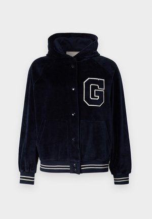 Sudadera de terciopelo de color marino con cierre de botones en la parte delantera, un gran parche de la letra "G" en blanco, dos bolsillos laterales y puños y dobladillo de canalé a rayas.