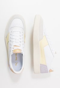 Białe i beżowe trampki z teksturowaną kremową podeszwą, wykonane z materiałów skórzanych i zamszowych, z złotymi akcentami i perforowanymi detalami na palcach.