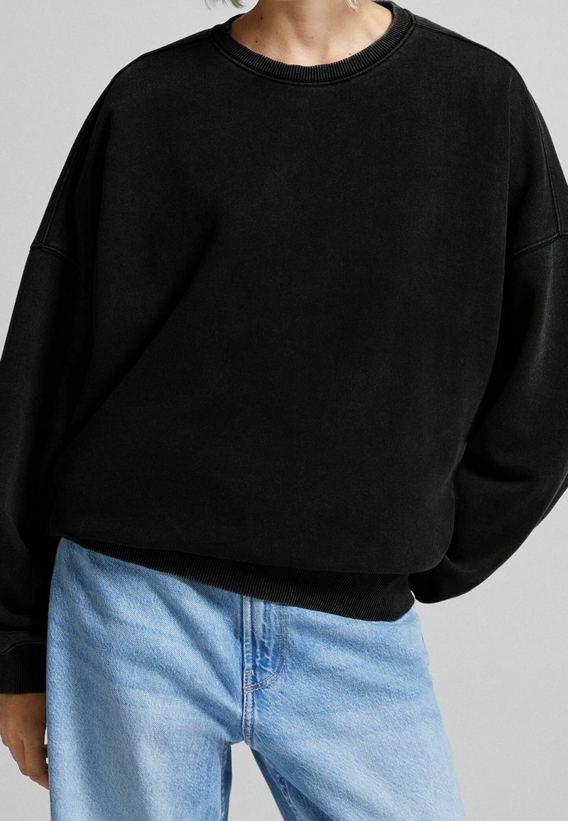 Sudadera negra confeccionada en tejido suave, con hombros caídos, cuello y puños de canalé, y un corte relajado, combinada con jeans de un color azul claro.