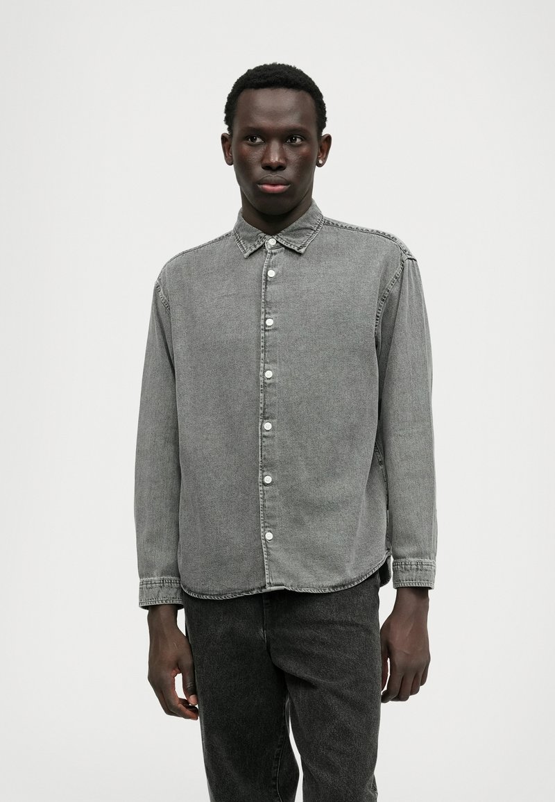 Camisa de denim cinza com colarinho tipo botão, fecho frontal com botões, mangas longas e bainha arredondada. Usada com calças de denim cinza a combinar.