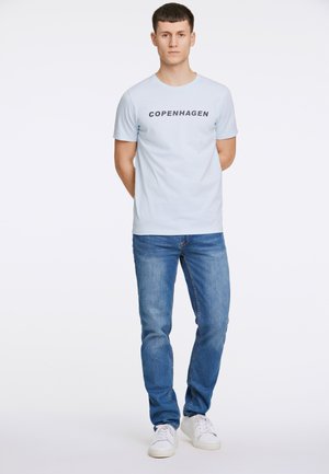 Lichtblauw katoenen T-shirt met "COPENHAGEN" in het zwart gedrukt, gecombineerd met blauwe denim jeans. Model staat met handen achter de rug.