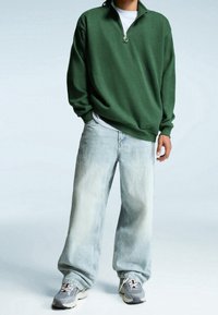 Person wearing a green quarter-zip sweatshirt, white undershirt, light wash loose jeans, and gray sneakers standing against a light background.