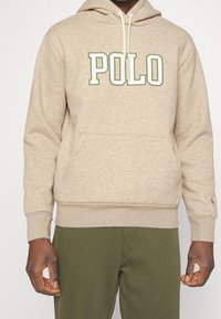 Beige mikina s kapsou na klokaní, kapucí se šňůrkou a logem "POLO" v zelené a bílé barvě na přední straně. Dlouhé rukávy s vroubkovanými manžetami.