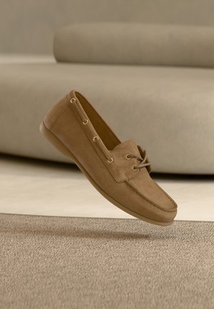 Chaussure en daim marron à enfiler avec œillets dorés et accents de lacets. Bout arrondi et semelle plate. Sur un fond beige texturé.