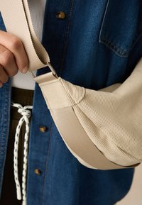 Bēša plecu soma no teksturēta auduma ar platu siksnu un metāla sprādzi, nēsāta pār džinsa jaku. Redzamas šuvju vietas un dizaina detaļas.