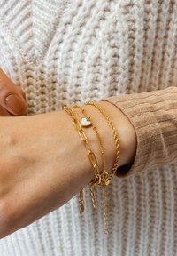 Pulseras de cadena de oro superpuestas en una muñeca, con un acento de corazón y diferentes estilos de eslabones. El fondo muestra un suéter de punto texturizado.