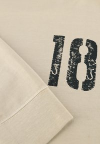 Cremfarbener T-Shirt mit einem abgenutzten schwarzen "18" Druck. Weicher, strukturierter Baumwollstoff mit Rundhalsausschnitt und genähten Ärmelabschlüssen.