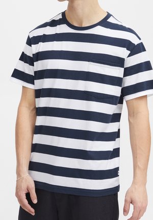 Mężczyzna w krótkim rękawie z granatowo-białymi poziomymi paskami, z kieszenią na piersi i czarnymi spodniami na białym tle.