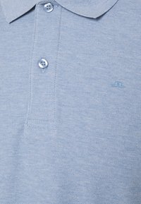 Polo bleu clair texturé avec deux boutons visibles et un petit logo brodé sur le côté droit de la poitrine.