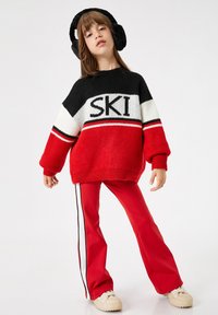 Roter und schwarzer Strickpullover mit dem Text "SKI", kombiniert mit roten Hosen mit seitlichen Streifen und klobigen beigen Schuhen. Strukturiert, oversized Passform.