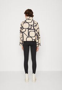 Giacca beige con stampa geometrica nera, design cropped, maniche lunghe e colletto alto, abbinata a leggings neri e stivaletti bianchi alla caviglia.