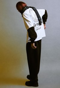 T-shirt bianca oversize con maniche nere e stampa grafica, abbinata a pantaloni neri larghi e a una sciarpa a quadri scuri, il tutto su uno sfondo grigio.