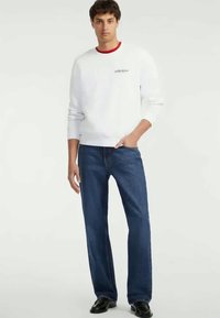 Sweatshirt branca com detalhe vermelho na gola, apresentando um pequeno logotipo. Jeans azul-escuro com corte direito. Sapatos pretos. Conjunto casual.