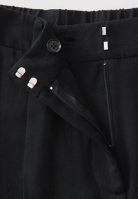 Ceinture d'un pantalon noir avec fermeture éclair ouverte, fermeture à bouton, deux agrafes métalliques à œil et crochet, et élastique au dos pour plus de flexibilité.