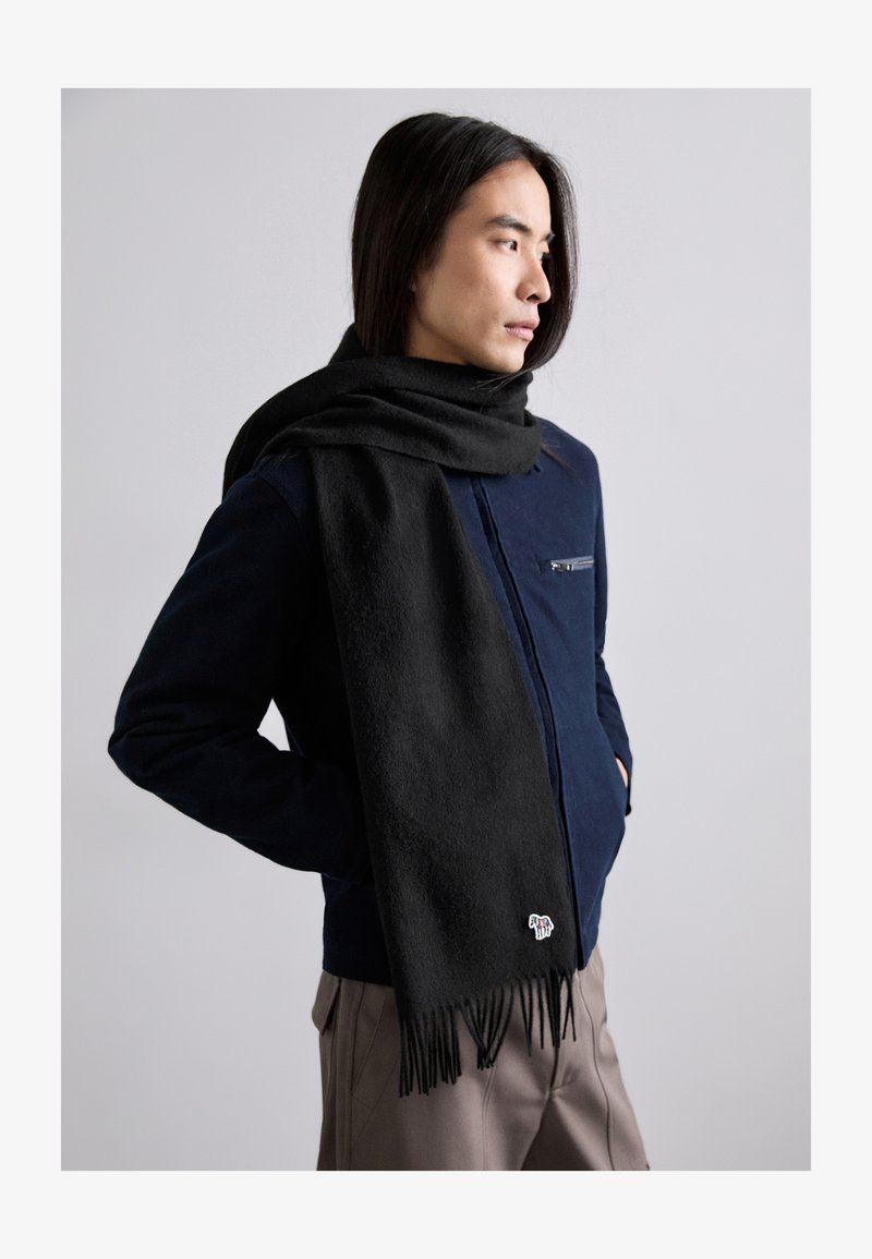 Écharpe en laine noire avec des franges, posée sur une veste bleu foncé, dotée d'une poche zippée et d'un petit détail brodé.