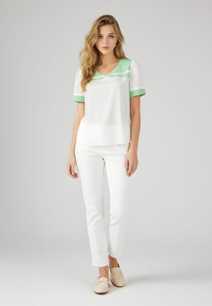 Mujer joven con largo cabello rubio que lleva pantalones blancos, mocasines crema y una blusa blanca con cuello y puños verdes, de pie frente a un fondo liso.