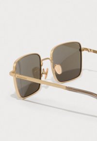 Τετράγωνα γυαλιά ηλίου με χρυσό μεταλλικό σκελετό, σκούρους φακούς και έντονη λεπτομέρεια στους βραχίονες. Χαραγμένο λογότυπο στο πλάι.