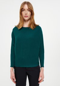 Teal geribbelde trui met een boothals en lange mouwen, gecombineerd met zwarte broek. De stof lijkt zacht en textuur te hebben.