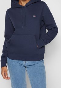 Sudadera de algodón azul marino con bolsillo tipo canguro, capucha con cordón y un pequeño logotipo en el pecho. Textura suave, corte holgado.