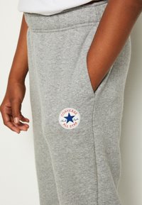 Grå sweatpants lavet af blødt stof, med sidelommer og et rundt broderet Converse-logo på venstre lår.
