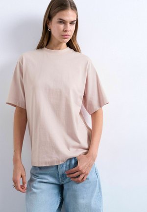 Jeune femme aux longs cheveux portant un t-shirt ample rose pâle et un jean bleu clair, debout contre un mur blanc.