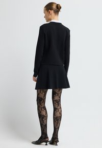 Mustat neuleet, joissa on tiivis siluetti, kerroksittain leveästi alas lähtevän mustan hameen päällä. Läpinäkyvät mustat sukkahousut kukkakuvioilla ja matalapohjaiset mustat kengät.