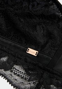 Tela de encaje negra con patrón floral y bordes festoneados, que presenta una pequeña etiqueta metálica grabada con la palabra "Rosie."