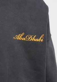 Ciemnoszary sweter z napisem "AbuDhabi" w pomarańczowym kursywie. Miękka faktura, okrągły dekolt i długie rękawy. Prosty design.