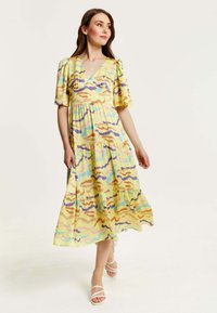 Maxi dress in yellow fabric featuring a colorful abstract pattern. V-neck, puff sleeves, and tiered design. Worn with white strappy sandals.