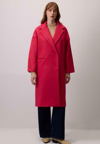 Rózsaszín, dupla soros gyapjú kabát széles hajtókákkal, túlméretezett fazonnal, hosszú ujjakkal és két elülső zsebbel. Beigazított bézs pulóver és sötét farmernadrág felett viselve.