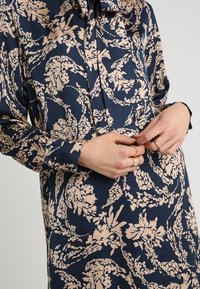 Marinblus med blommönster i ljus persika, med långa ärmar, knäppningar vid ärmsluten och en fluga vid halsringningen. Slät satintextur.