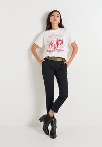 T-shirt blanc avec un design graphique rouge, représentant des montagnes et des cactus. Assorti avec un pantalon noir ajusté et des bottines noires à talons.