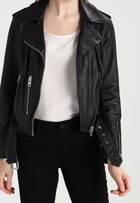 Schwarze Lederbikerjacke mit silbernen Reißverschlüssen, mit einem geschlitzten Kragen und seitlichen Schnallen. Getragen über einem weißen Oberteil und schwarzen Jeans.