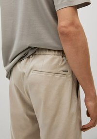 Gros plan d'une personne portant un pantalon beige avec une ceinture élastique et une poche arrière, associé à une chemise gris clair à manches courtes.