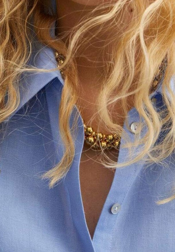 Niebieska koszula na guziki z krótkimi rękawami i kołnierzykiem. Przy dekolcie znajduje się ozdobny naszyjnik z złotych koralików. Widoczne kręcone blond włosy.