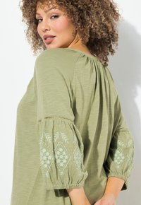 Oliwkowa bluzka z rękawami o długości trzech czwartych, z bufiastymi rękawami, zdobiona jasnoniebieskimi haftami w kwiaty i subtelną teksturą. Luźny fason z marszczonym tyłem.