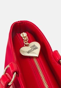 Czerwona torebka syntetyczna z ozdobnym złotym suwakiem oraz zawieszką w kształcie serca z napisem "LOVE MOSCHINO". Gładka tekstura z metalowymi akcentami.